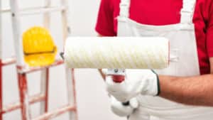 Caucasian house painter worker in white work overalls and with protective gloves, he holds the roller for painting in hand. Construction industry. Work tools.